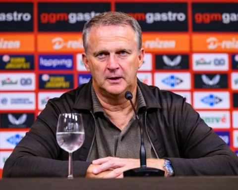 John van den Brom zit achter een microfoon tijdens een persmoment, met sponsorwand op de achtergrond.