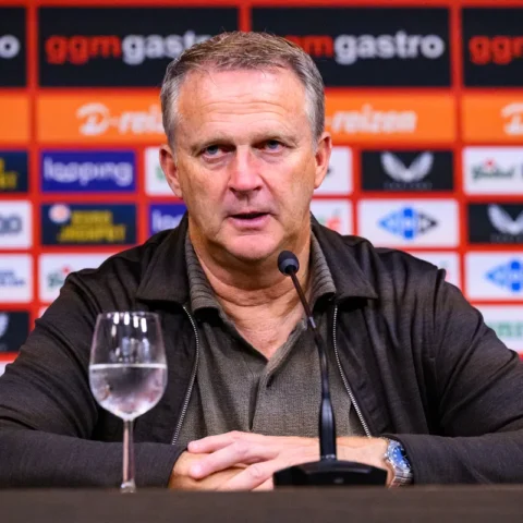 John van den Brom zit achter een microfoon tijdens een persmoment, met sponsorwand op de achtergrond.