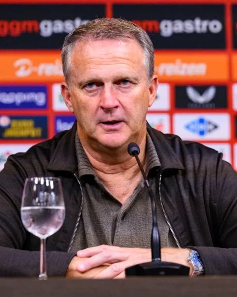 John van den Brom zit achter een microfoon tijdens een persmoment, met sponsorwand op de achtergrond.